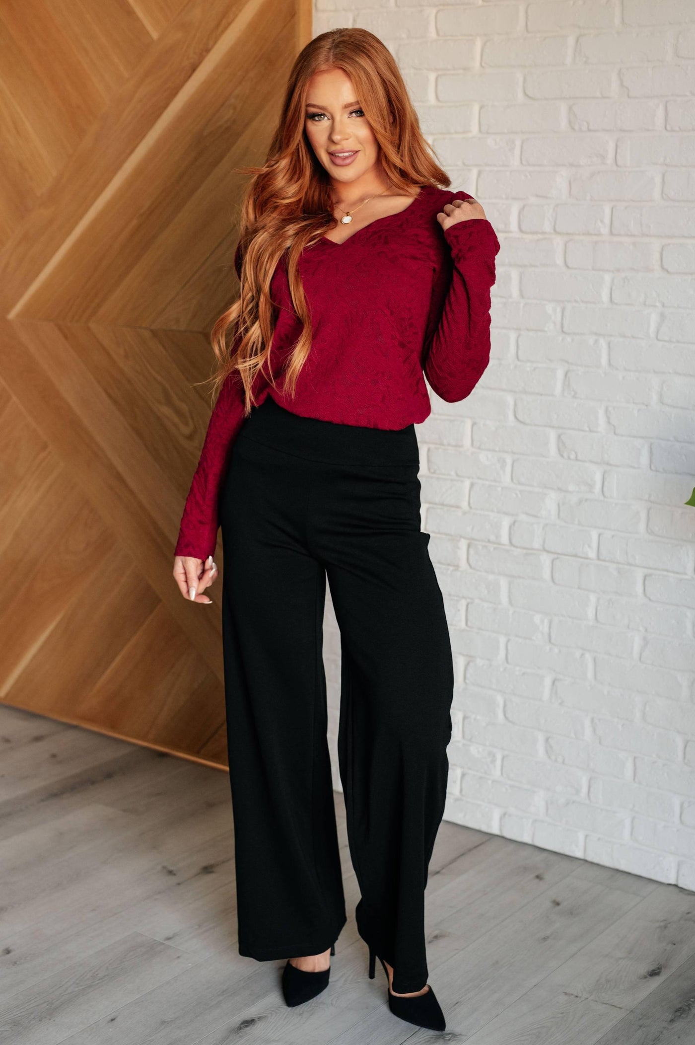 Model in red ruched sleeve top and black pants, standing indoors.