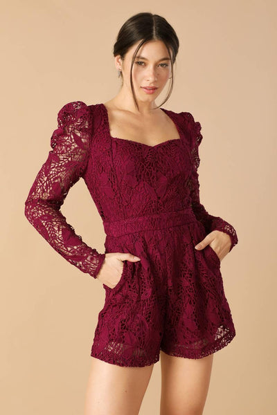 Model wearing burgundy lace romper with hands in pockets.