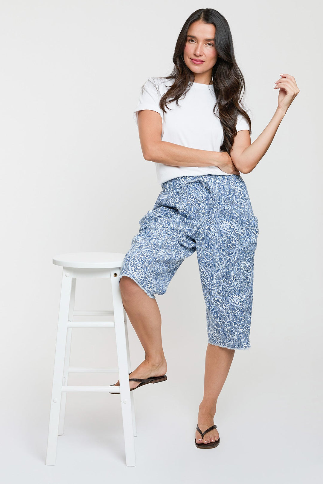 Woman wearing a white t-shirt and blue patterned shorts standing on a white stool against a white background